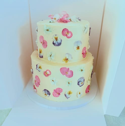 A customer's two-tier wedding cake decorated with colorful freeze-dried BEGONIA edible flowers, shown inside a white cake box.