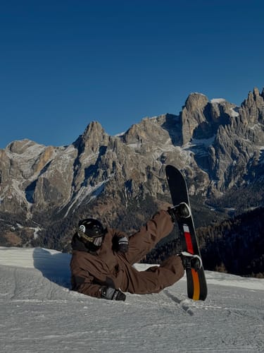 Reviewer in baggy brown snow pants, sitting playfully in the snow with mountains in the background.