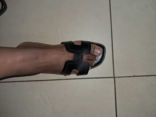 A customer's foot seen from above wearing the black Miss Black Syrup 2 Sandal on a tiled floor.