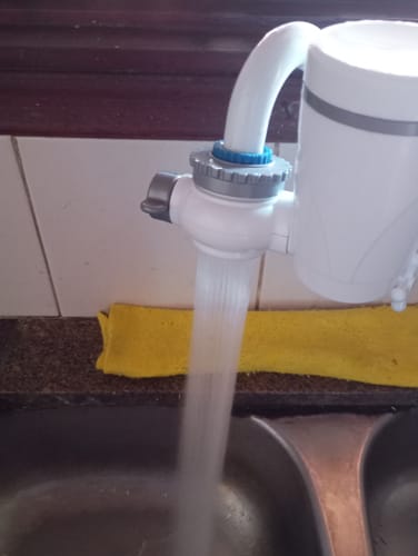 Cliente muestra su Filtro Purificador de Agua blanco instalado en un grifo de cocina, con el agua fluyendo hacia el fregadero.