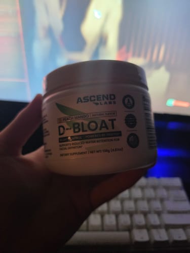 Customer holding a white tub of D-BLOAT powder in Peach Mango flavor in their hand, with a desk and keyboard in the background.