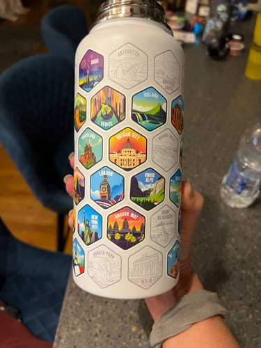 Customer holding the white Lets Wander™ World Explorer Water Bottle with colorful travel stickers applied over some engraved outlines.