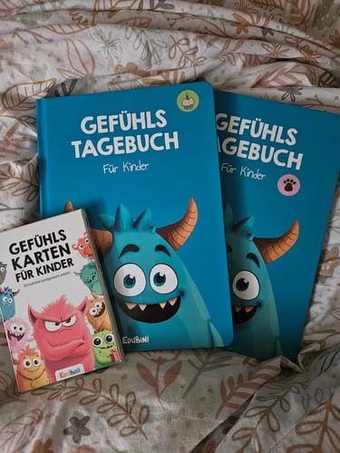 Ein Kunde zeigt die Edubini Gefühlskarten und zwei blaue Gefühlstagebücher auf einer gemusterten Decke.