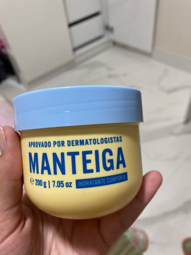 Cliente segurando o pote amarelo da Manteiga Hidratante com tampa azul claro, em um ambiente interno.