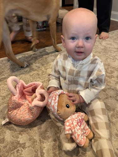 Customer's baby sitting on a rug, holding the soft, tanned Personalized Pudding Baby Girl doll with its matching pink bag nearby.
