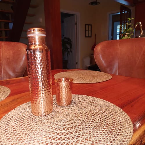 Customer's hand-hammered Pure Copper Water Bottle and a matching cup on a woven placemat on a table.
