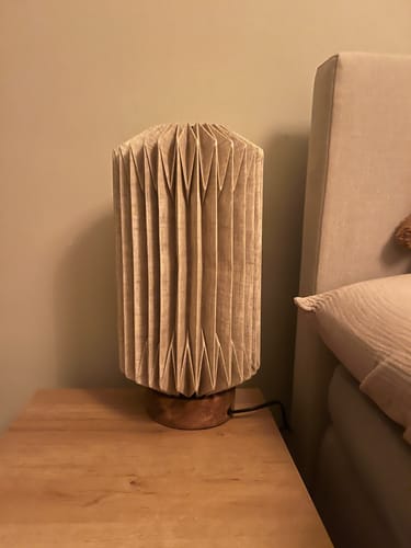 Recensent toont de onverlichte Linen Tafellamp met geplooide kap op een houten nachtkastje.