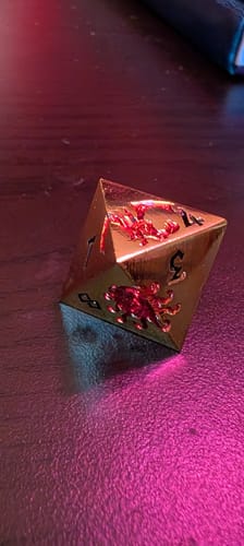 Close-up of a customer's gold and red metal Monster Manual die on a dark surface under pink light.