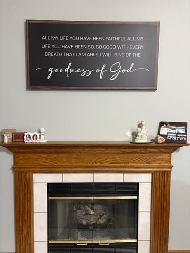 Reviewer's Goodness Of God Faith Sign II hanging on a gray wall above a wooden fireplace.