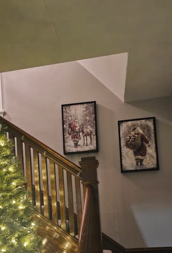 Customer's two framed Vintage Christmas Santa Claus canvases hanging on a wall by a wooden staircase.