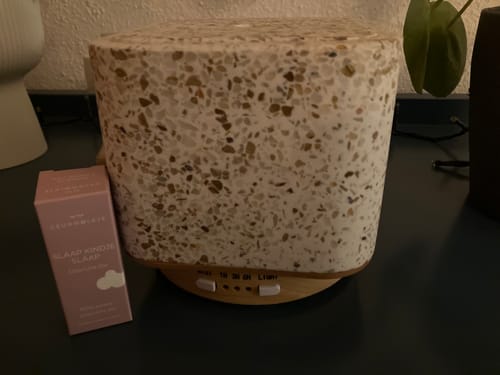 Recensent toont de verpakking van de 'Slaap kindje slaap' olie naast een lichte terrazzo aromadiffuser.