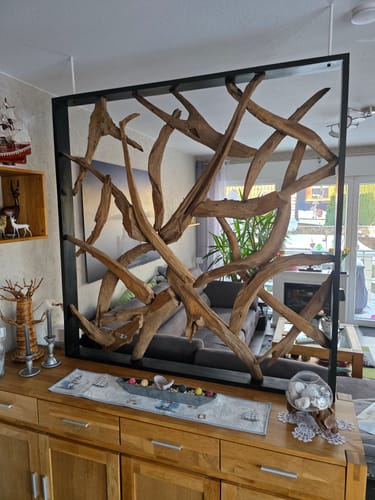 Kunde zeigt den MAZE Raumteiler aus Teakholz, der dekorativ auf einer Anrichte aus Holz im Wohnzimmer steht.
