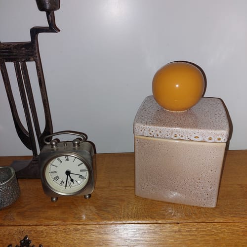 Recensent toont de gespikkelde Runa pot met een oranje bol als handvat, staand op een houten meubel.