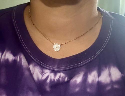 Customer wearing the White Blossom Flower Necklace. The gold beaded chain rests on their neck against a purple shirt.