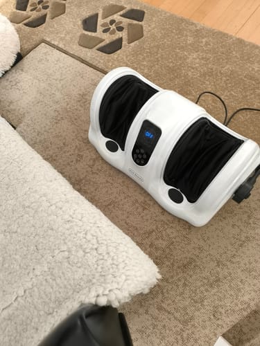 Anmelders hvide fod- og lægmassageapparat står på et gulvtæppe. Displayet er tændt.