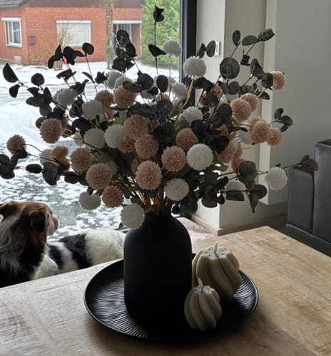 Klant toont een vol, gemengd boeket met witte en lichtroze Kiku bloemen en eucalyptus in een zwarte vaas.