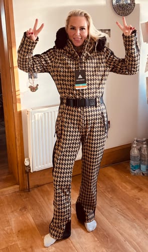 Reviewer posing indoors in the slim-fit tan houndstooth one-piece ski suit with its black faux fur hood.