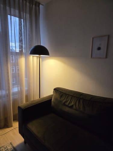 Recensent toont de zwarte by fonQ Bloop vloerlamp die verlicht is in een woonkamer naast een donkere bank.