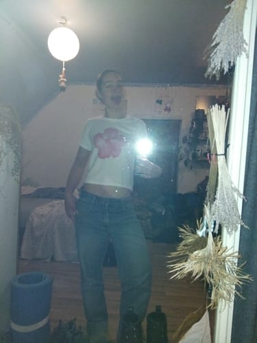 Reviewer in a mirror selfie wearing the white hibiscus baby tee and blue jeans in a bedroom.