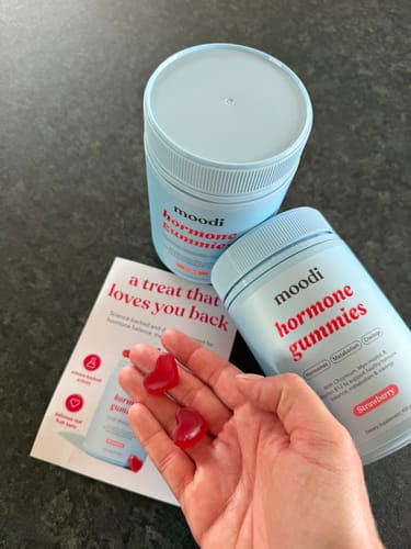 Reviewer's hand holding two red, heart-shaped Hormone Gummies in front of the two bottles from the bundle.