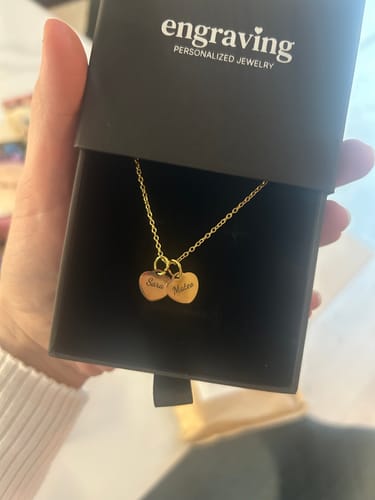 Customer holding a gold Heart Charm Necklace with two engraved charms inside its jewelry box.