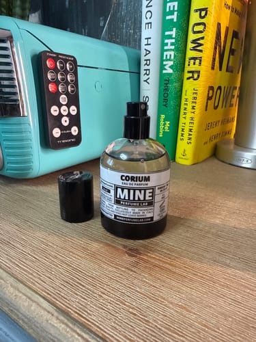 Recensore mostra la sua boccetta di profumo CORIUM su un mobile in legno, con il tappo accanto.