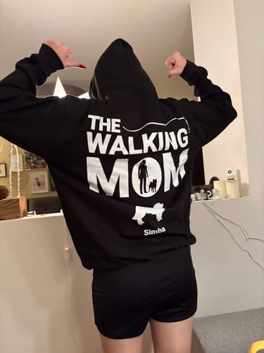 Kundin zeigt die Rückseite ihres schwarzen personalisierten Hoodies mit dem Aufdruck "THE WALKING MOM", Hundegrafiken und dem Namen "Simba".