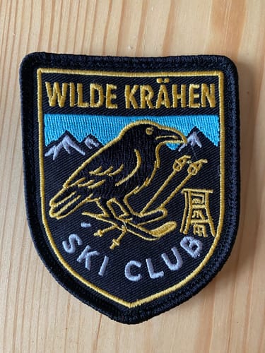 Rezensent zeigt einen individuell gefertigten Patch für den 'Wilde Krähen Ski Club' in Nahaufnahme auf einer Holzoberfläche.