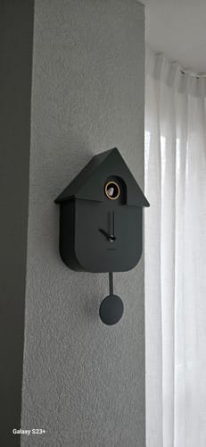 Recensent toont de donkere, moderne Cuckoo wandklok zonder cijfers, hangend aan een grijze muur.