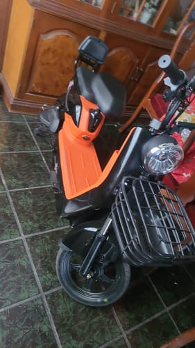 Un cliente muestra su bicicleta eléctrica Tori 800 negra con naranja, estacionada en el interior, mostrando la cesta delantera.