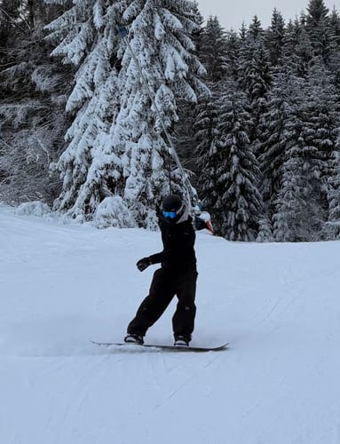 Reviewer wearing baggy dark Snow Pants while snowboarding on a snowy slope in front of a forest.