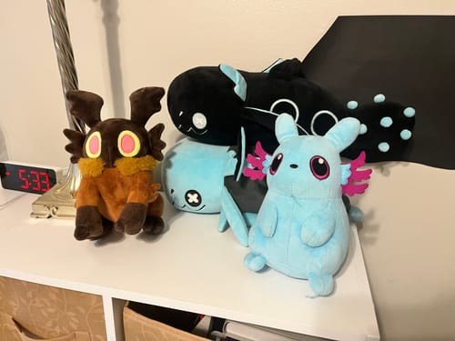 Customer's black Glowing Cyan Lizard Plush sitting on a white shelf with other colorful plush toys.
