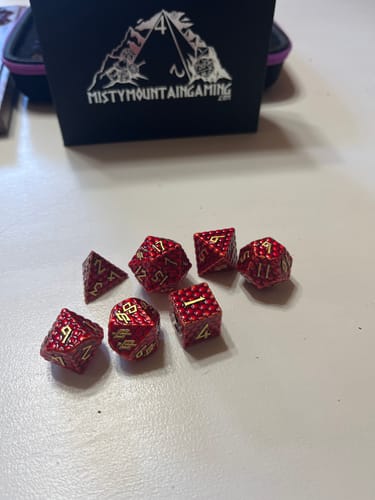 Reviewer's Red Dragon's Egg Metal Dice Set with textured red finish and gold numbers spread on a table in front of its box.