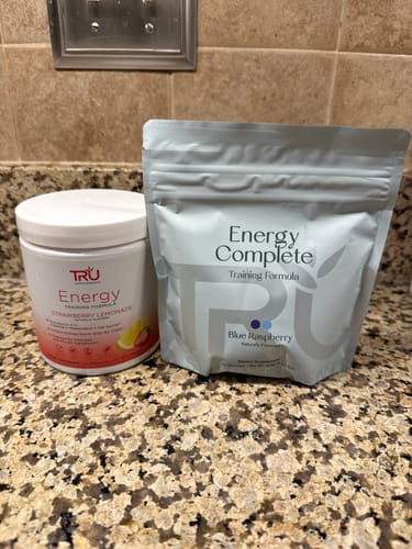 Reviewer shows the new bag packaging for Energy Complete next to the previous plastic container on a counter.