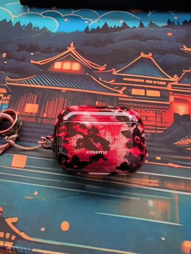 Reviewer's glossy Obsession AirPods Case with a red and black pattern, shown on a desk mat.