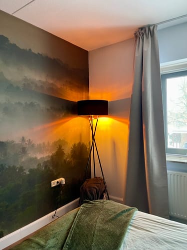 Beoordelaar toont de zwarte Rivel Vloerlamp in een hoek. De lamp is aan en geeft een warm, sfeervol licht op de muur.