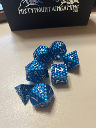 Customer's blue, textured Dragon's Egg Metal Dice Set scattered on a table in front of the product's box.