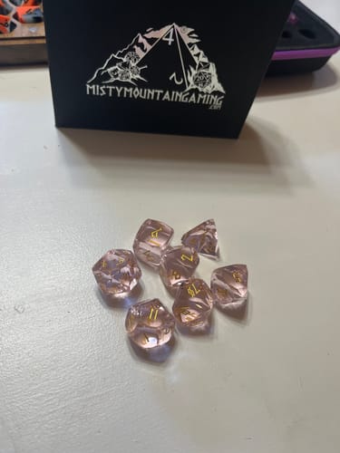 A customer's Baby Pink Topaz Multifaceted Glass Dice Set scattered on a table in front of its black storage box.
