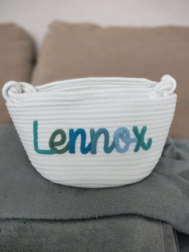 Customer's personalized white knitted basket with the name 'Lennox' in shades of blue and green yarn.