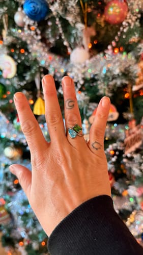 Customer's hand wearing the Navae Birthstone Ring with green and light blue stones in front of a Christmas tree.