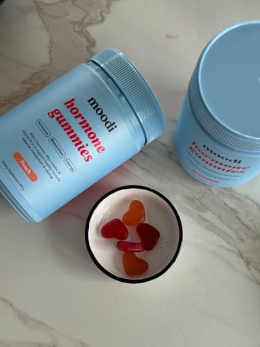 Reviewer's light blue container of Peach Hormone Gummies with several red, heart-shaped gummies in a small bowl.