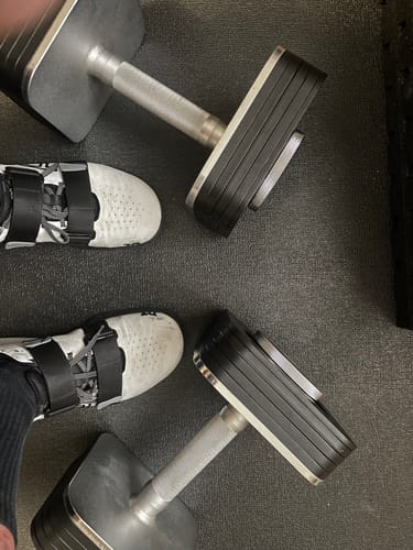 Customer's two Strength Shop Adjustable Dumbbells on a black floor mat, viewed from above next to their feet in lifting shoes.
