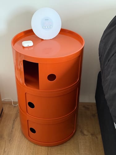 Recensent gebruikt de oranje Kartell Componibili Kast als een handig nachtkastje naast een bed.