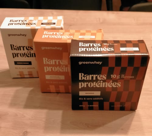 Avis d'un client montrant trois boîtes de barres protéinées Greenwhey sur une table en bois, saveurs brownie et cookie dough.
