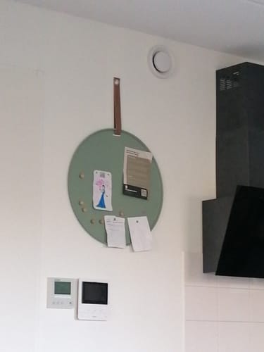 Recensent toont het ronde, groene Perky memobord hangend aan een muur met briefjes en tekeningen erop.