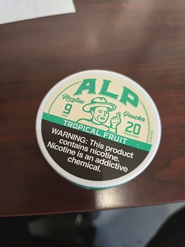 Customer's round tin of ALP Tropical Fruit Nicotine Pouches sitting on a dark wooden desk.