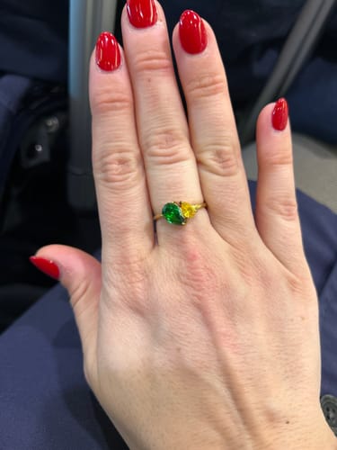 Reviewer's hand, with red nails, wearing the gold Leesha Birthstone Ring with one green and one yellow pear-cut stone.