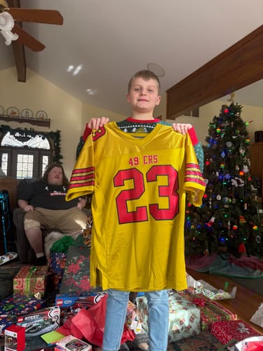 A person holds up a custom old gold football jersey with red lettering and the number 23 in a living room.