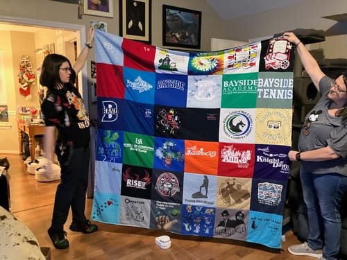 Customer and another person holding up their large, colorful Premium T Shirt Quilt in a living room.