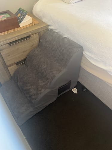 Customer's grey Foam Pet Steps placed on a dark carpet next to a bed and a wooden nightstand.